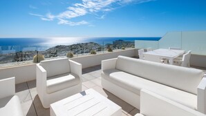 A rooftop terrace with white furniture, offering a view of the sea and a coastal town.