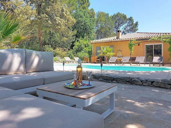 A poolside area with a white sofa, a wooden table with a bottle and glasses, and a pool with lounge chairs in the background.