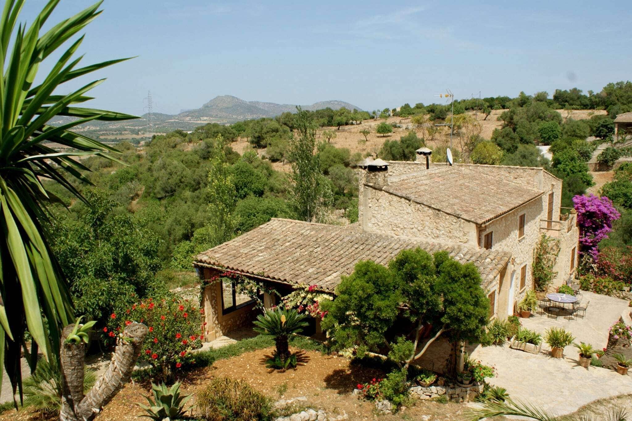 Un bâtiment en pierre, surmonté d’un toit en tuiles, entouré d’une végétation luxuriante et d’un jardin où poussent diverses plantes et fleurs.