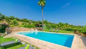 Una piscina rectangular con solárium de piedra, rodeada de césped y palmeras, bajo un cielo azul despejado.
