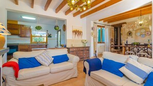 A spacious living area with two sofas, wooden beams, a chandelier, and a kitchen in the background.