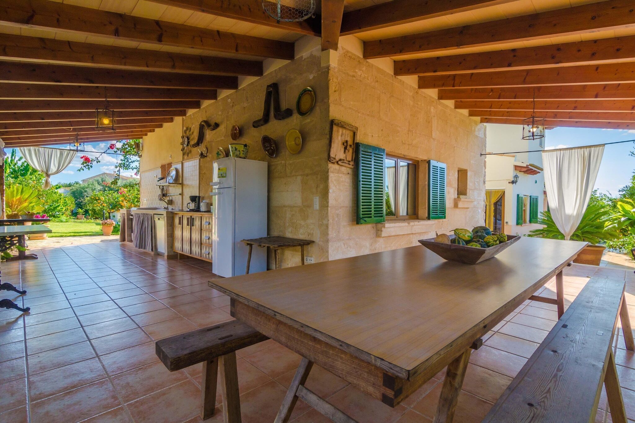 A spacious patio area with a long wooden table and benches, a kitchen area with a refrigerator and a sink, and a view of a lush garden.