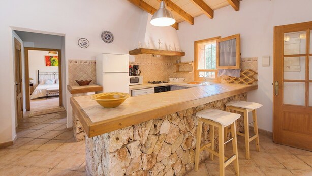 A kitchen with a stone island, wooden stools, a hanging light fixture, and a view of the adjacent bedroom.