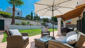 An outdoor patio area with wicker furniture, a large umbrella, and a view of a well-maintained garden and buildings in the background. - El Pinar - Villa With Private Pool. Free Wifi (Cala Sant Vicenç, Illes Balears)