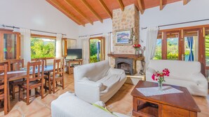 A spacious living area with a stone fireplace, wooden furniture, and large windows offering a view of greenery. - Can Reure (can Paris) - Villa With Private Pool (Inca, Illes Balears)