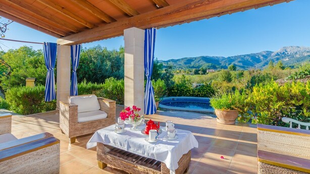An outdoor patio with a table set for tea, surrounded by wicker furniture and blue-striped curtains, overlooking a pool and lush greenery. - Can Reure (can Paris) - Villa With Private Pool (Inca, Illes Balears)