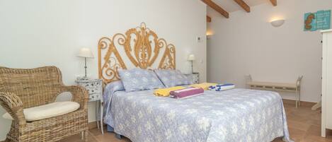 A bedroom with a bed, a wicker chair, a bedside table, and a bench.