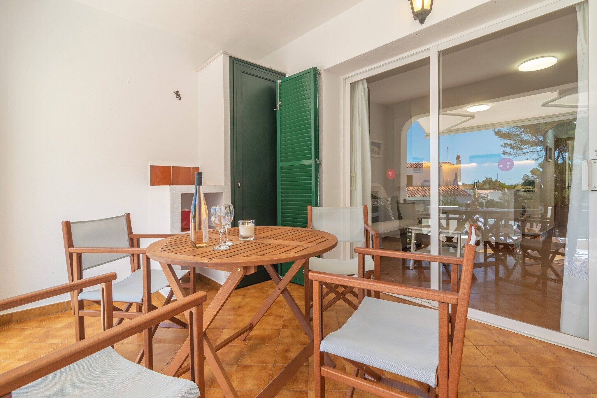 Een kamer met een houten tafel en stoelen, een groene deur en uitzicht door een glazen deur op een buitenruimte met tafels en stoelen.
