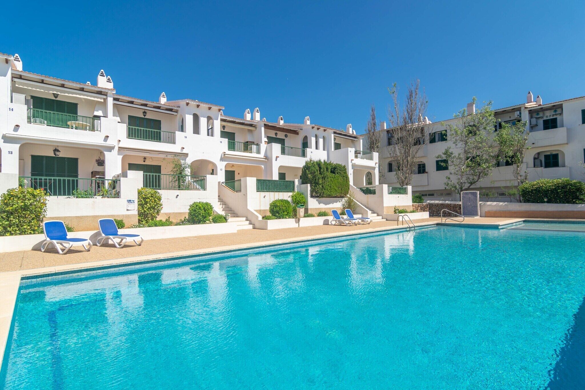A swimming pool with lounge chairs, a building with balconies, and a clear blue sky.