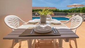 Une table à manger dressée pour quatre personnes, avec des chaises blanches, une plante en pot et une vue sur une piscine.