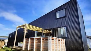 A modern two-story building with a dark exterior and a wooden pergola structure in the foreground. - “ VITAMIN SEA “ 
Beach ~ House  (Riverton / Aparima)