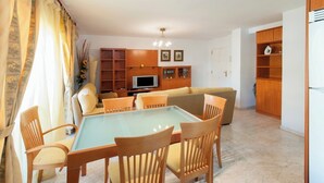 A living room with a glass-top dining table, wooden chairs, a sofa, a TV, wooden shelving, and a door leading to another room. - Cascades - Apartment In Dénia (Denia, Alicante)