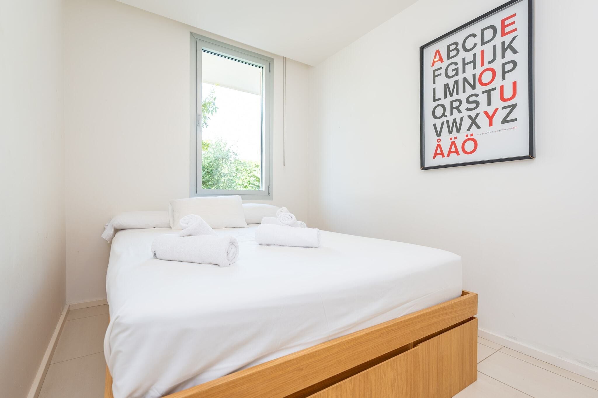 A neatly made bed with white linens and towels, a window with a view of greenery, and a wall-mounted alphabet poster.