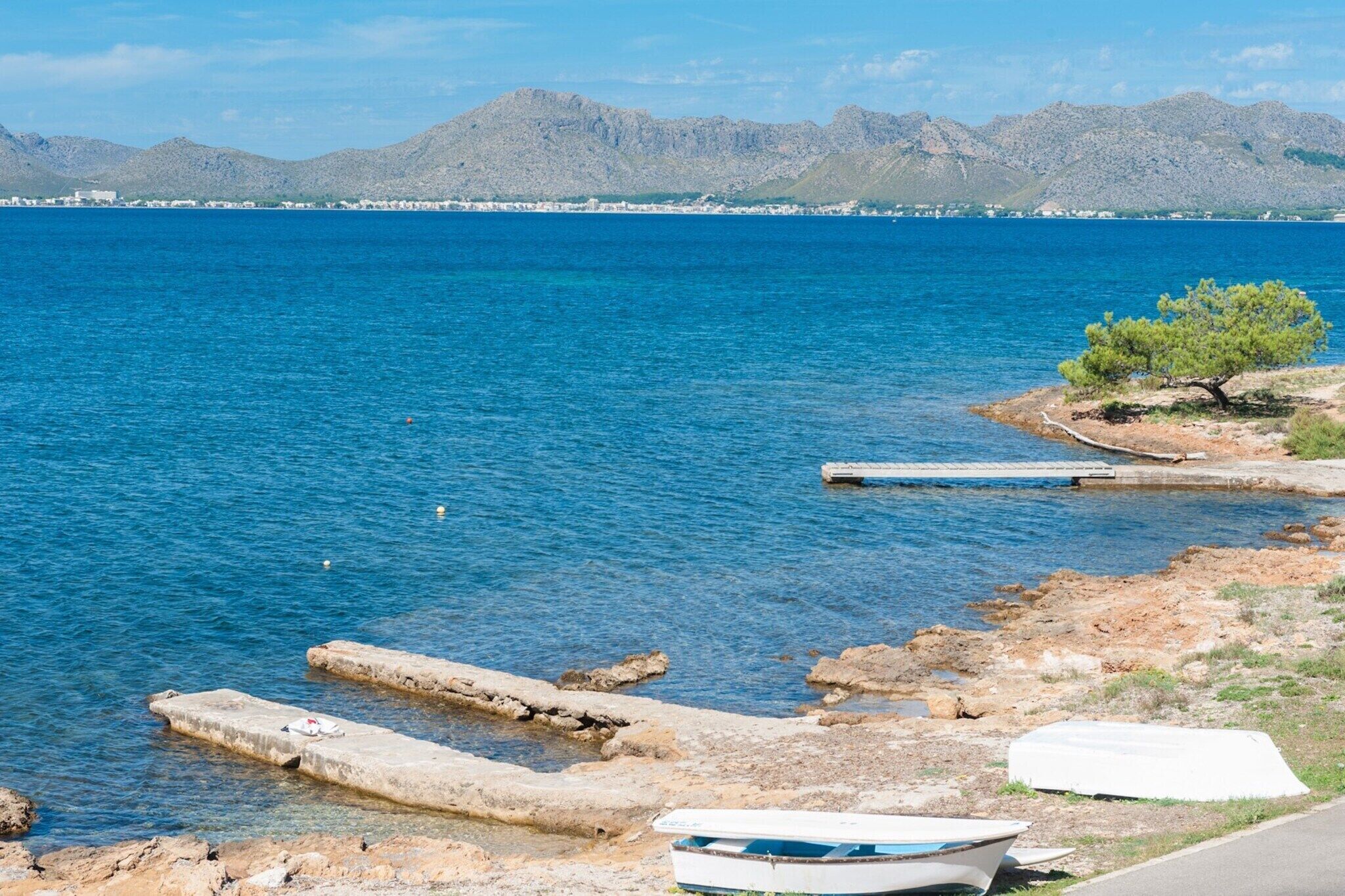 A coastal scene with a clear blue sea, rocky shore, a small boat, and distant mountains.