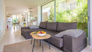 Een moderne woonkamer met een grijze bank, een ronde houten salontafel met een fruitschaal, en een groot raam met uitzicht op groen.