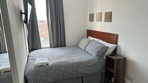 A neatly made bed with a grey and white striped bedspread and matching pillows, a wooden headboard, a small wooden nightstand with a remote control, and a view of a brick building through a window with grey curtains. - Home and Away (Eaglescliffe)