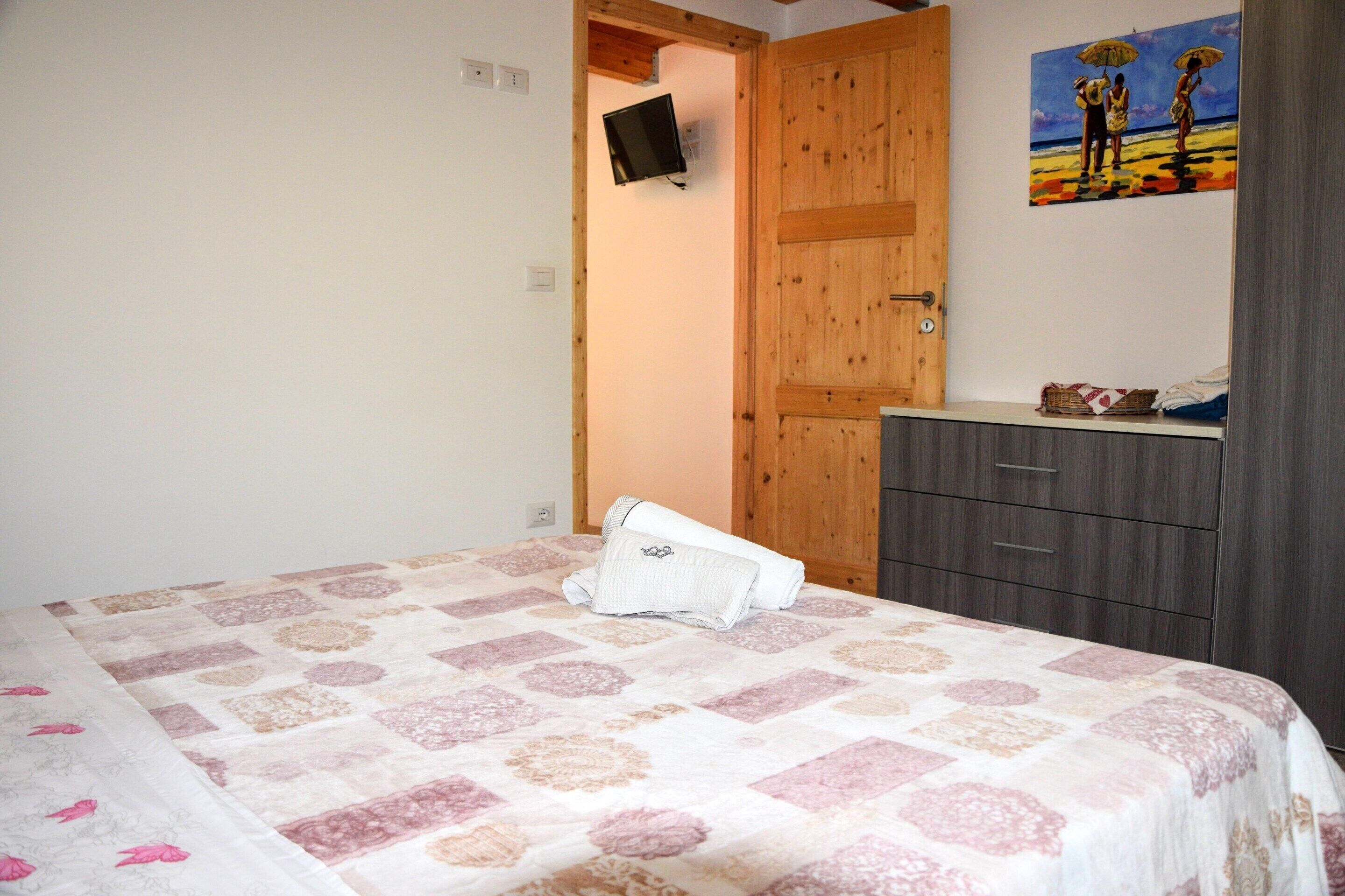 Ein Schlafzimmer mit einem Bett, einer Holztür, einem an der Wand montierten Fernseher und einem Gemälde über der Tür.