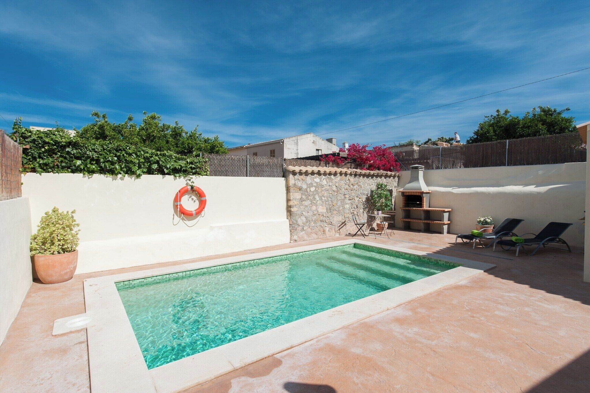 Une piscine rectangulaire à l’eau claire, entourée d’une terrasse en béton. On y trouve des chaises longues et une petite table avec des chaises, un pot de fleurs et un anneau de sauvetage fixé au mur.