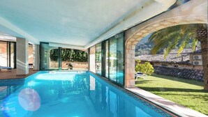 En swimmingpool med klart blåt vand, omgivet af store vinduer der giver udsigt over en have og en bygning med et tag af fliser.