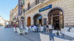 Un café al aire libre con mesas y sillas, un letrero que dice Bella Italia, y personas cenando.