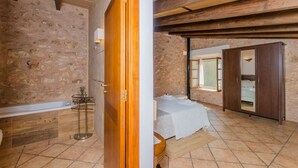 Una habitación con pared de piedra, vigas de madera en el techo, una cama con cubre cama blanco y un armario.