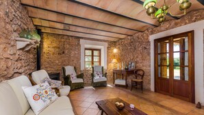 Una habitación rústica con paredes de piedra, techo de madera y una amplia puerta de madera. Hay dos sillones, un sofá y una mesa de madera con una cesta y un vaso.