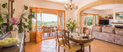 Un grand espace de vie comprenant une table à manger, des canapés et un téléviseur. Un balcon offre une vue sur la verdure.