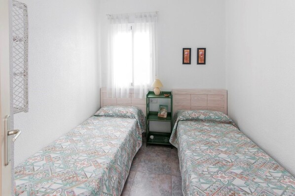 Una habitación pequeña y limpia con dos camas individuales, una ventana con cortinas traslúcidas, una mesita de noche con lámpara y dos cuadros en la pared.