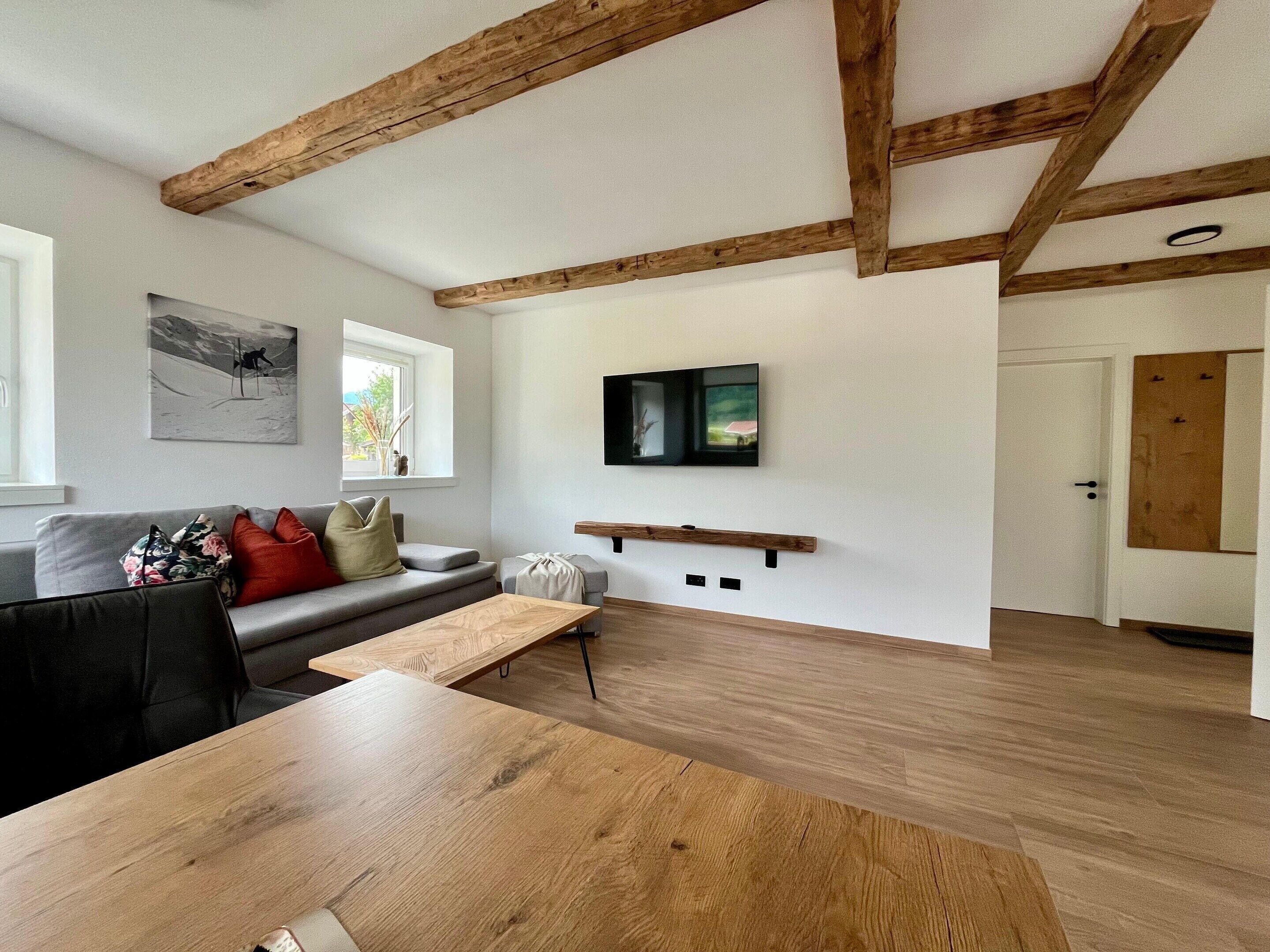 Ein modernes Wohnzimmer mit freiliegenden Holzbalken, einem grauen Sofa, einem Holztisch und einem flachbildschirmfernseher an der Wand.