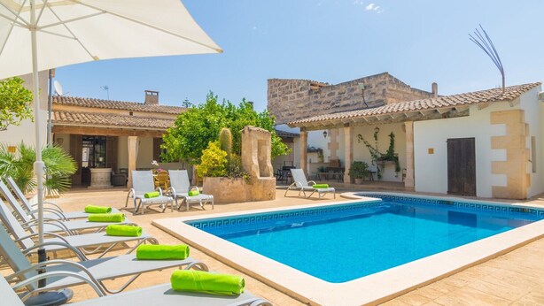Una piscina con sillas de descanso y sombrillas, rodeada de edificios con techos de tejas y vegetación.