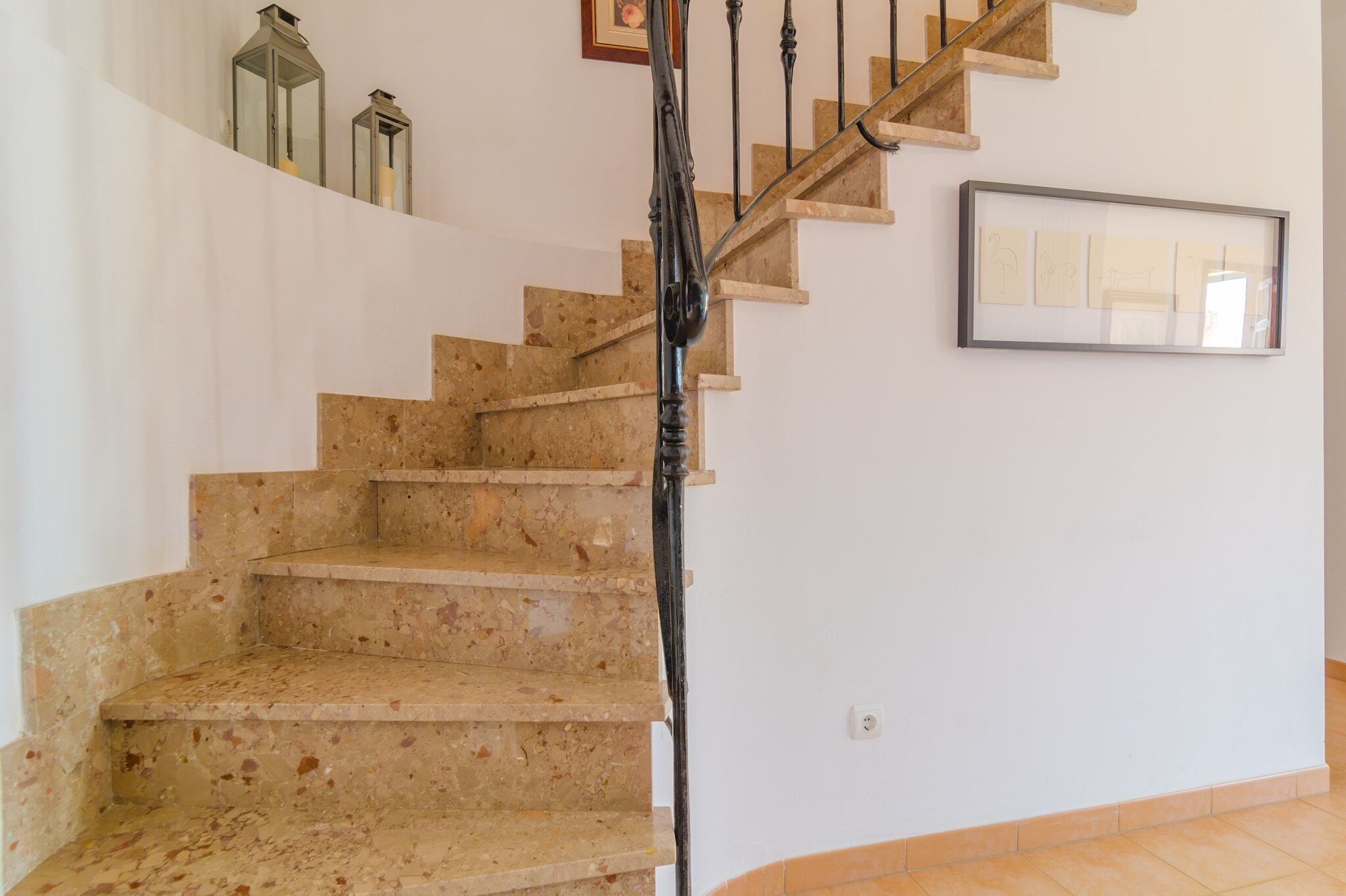 A staircase with a wooden handrail and beige steps, leading to a landing with a framed picture and two wall-mounted lanterns.