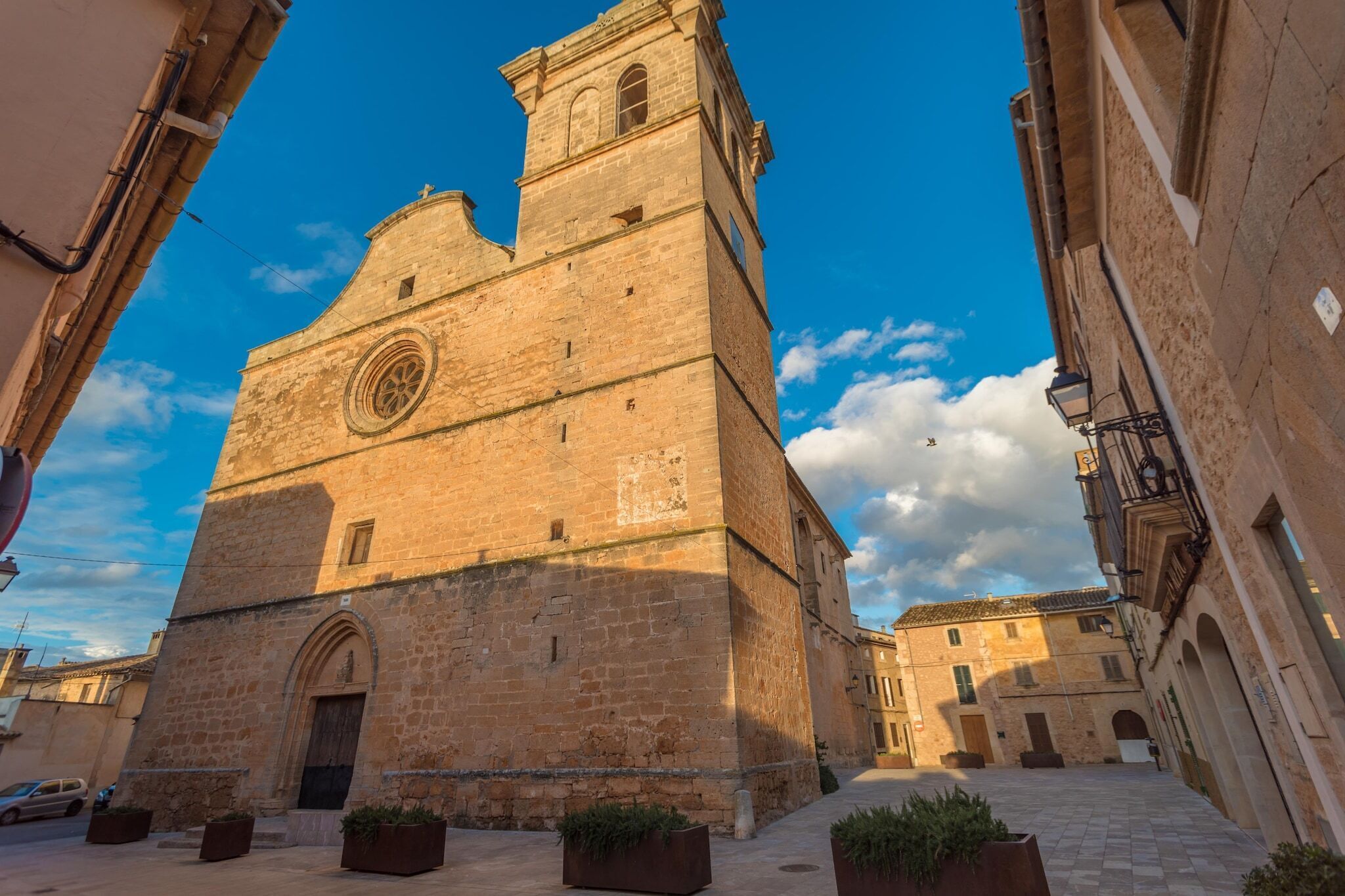 Un edificio histórico de piedra con una torre destacada y una entrada arqueada, rodeado de un patio empedrado y edificios colindantes.