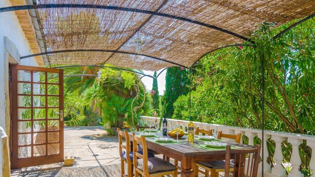 An outdoor patio with a wooden table and chairs, surrounded by lush greenery and a thatched roof. - Can Bolei - Villa With Private Pool In Sant Elm (Sant Elm (andratx), Illes Balears)