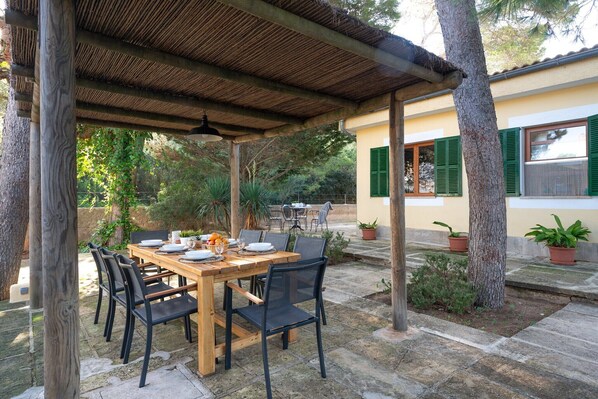 An outdoor patio with a wooden table and chairs, surrounded by stone tiles and potted plants.