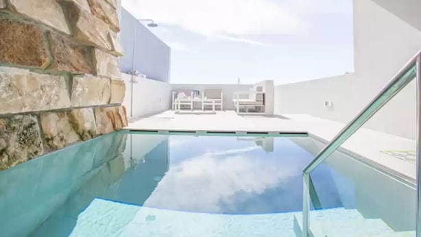 A modern indoor pool with clear glass walls, a stone wall on one side, and a view of the sky and outdoor seating area. - S'aigua 5 - Villa With Private Pool In S'illot (S'illot, Illes Balears)
