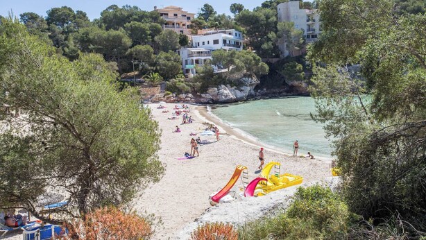 A beach with people sunbathing and swimming, a lifeguard tower, and buildings in the background. - Xalet S'escaleta - Villa With Private Pool (Cala Santanyí, Illes Balears)