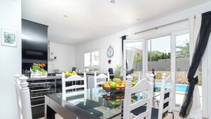 A dining area with a glass table, white chairs, and a fruit bowl. There is a kitchen area with a black cabinet and a TV. A pool is visible through the glass doors. - Xalet S'escaleta - Villa With Private Pool (Cala Santanyí, Illes Balears)