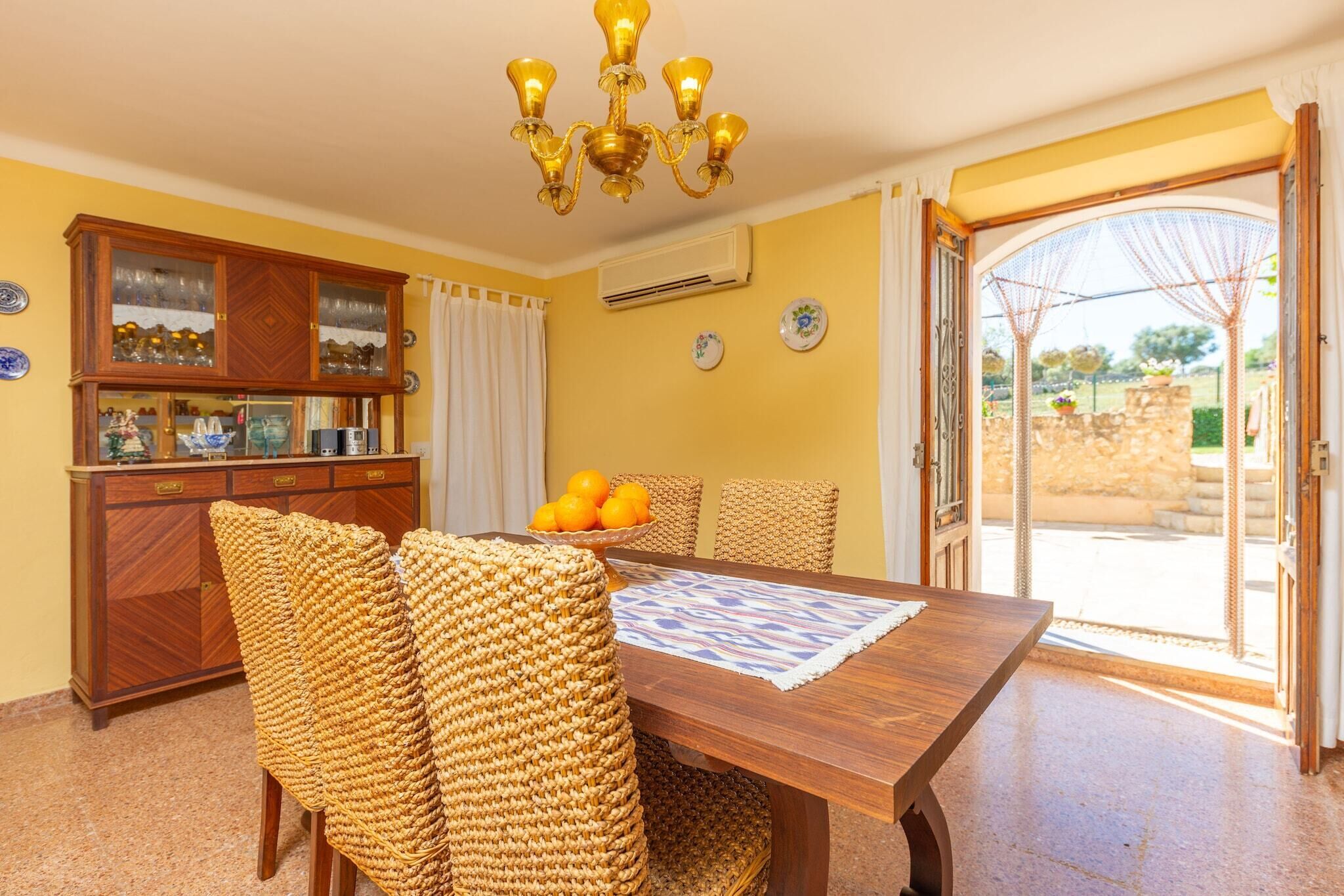A dining area with a wooden table, woven chairs, a chandelier, and a view through an open door to an outdoor area.