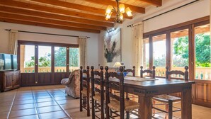 A dining area with a wooden table and chairs, a chandelier, and large windows with a view of greenery. - Ca'n Berguins - Villa With Private Pool In Búger (Buger, Illes Balears)