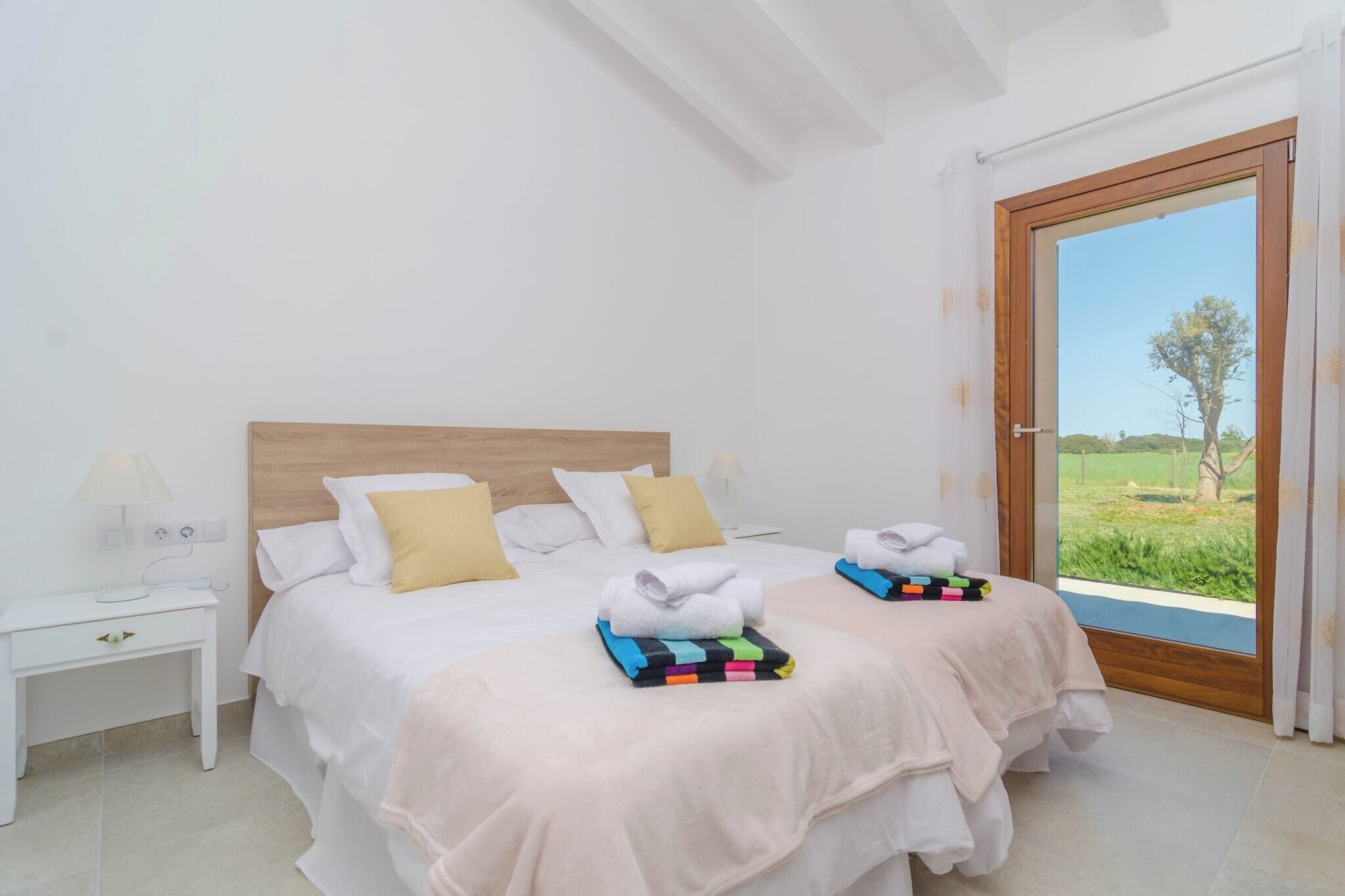 A double bed with white and yellow pillows, a nightstand with a lamp, and a view of a green landscape through a large window.