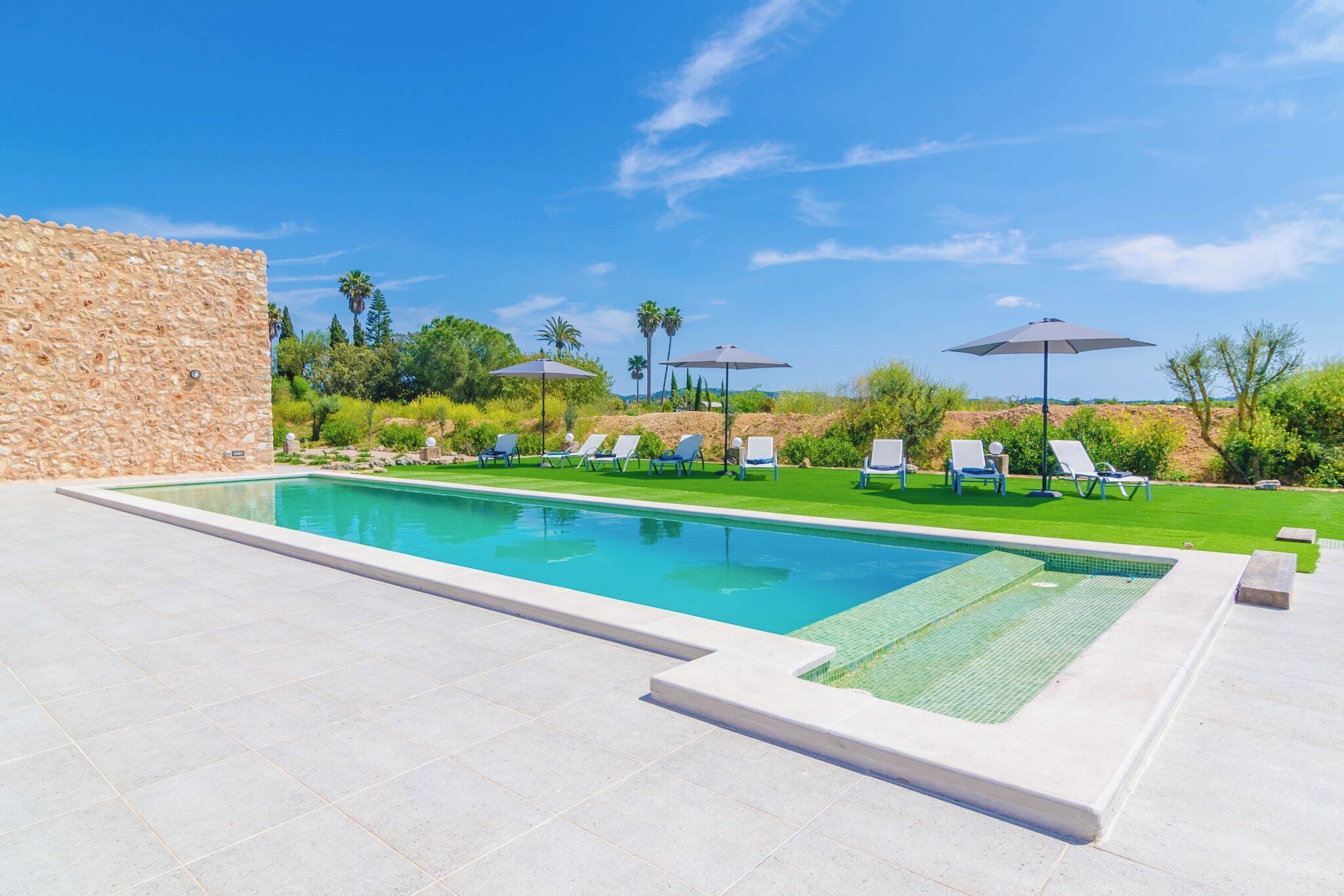 A swimming pool with lounge chairs and umbrellas, surrounded by greenery and a stone wall.