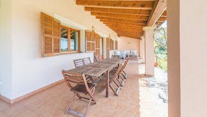 Un espace extérieur abrité, aménagé pour les repas, avec des tables et des chaises en bois, de grandes fenêtres équipées de volets et une vue sur l’extérieur.