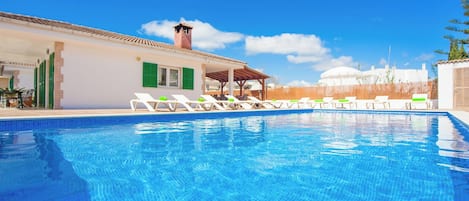 Ein Schwimmbecken mit Liegestühlen, einer überdachten Sitzgelegenheit und einem Gebäude mit einem Ziegeldach.