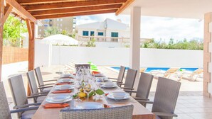 Een terras met een lange tafel gedekt voor een maaltijd, omringd door rieten stoelen, onder een houten pergola. Achtergrond: een zwembad.