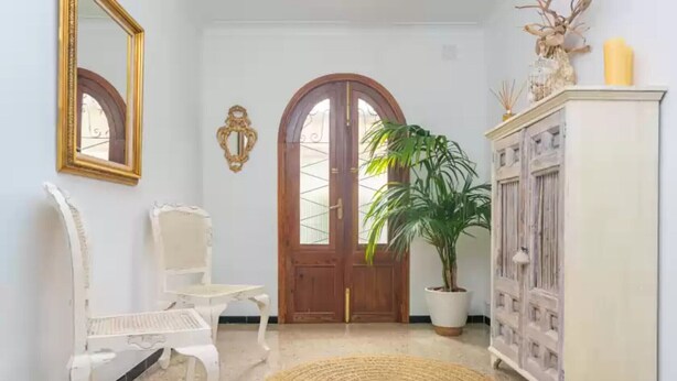 A hallway with a white chair, a round woven rug, a wooden door, and a cabinet with decorative items.