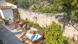 A sunlit patio with a wooden table and lounge chairs, surrounded by potted plants and flowering shrubs.