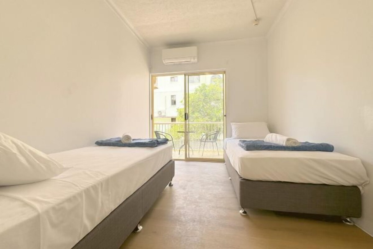 Una habitación pequeña y limpia con dos camas, una puerta corrediza de vidrio a un balcón y un equipo de aire acondicionado blanco en la pared.