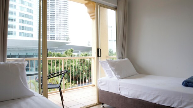 A hotel room with two beds, a balcony with a chair, and a view of a tall building and palm trees.