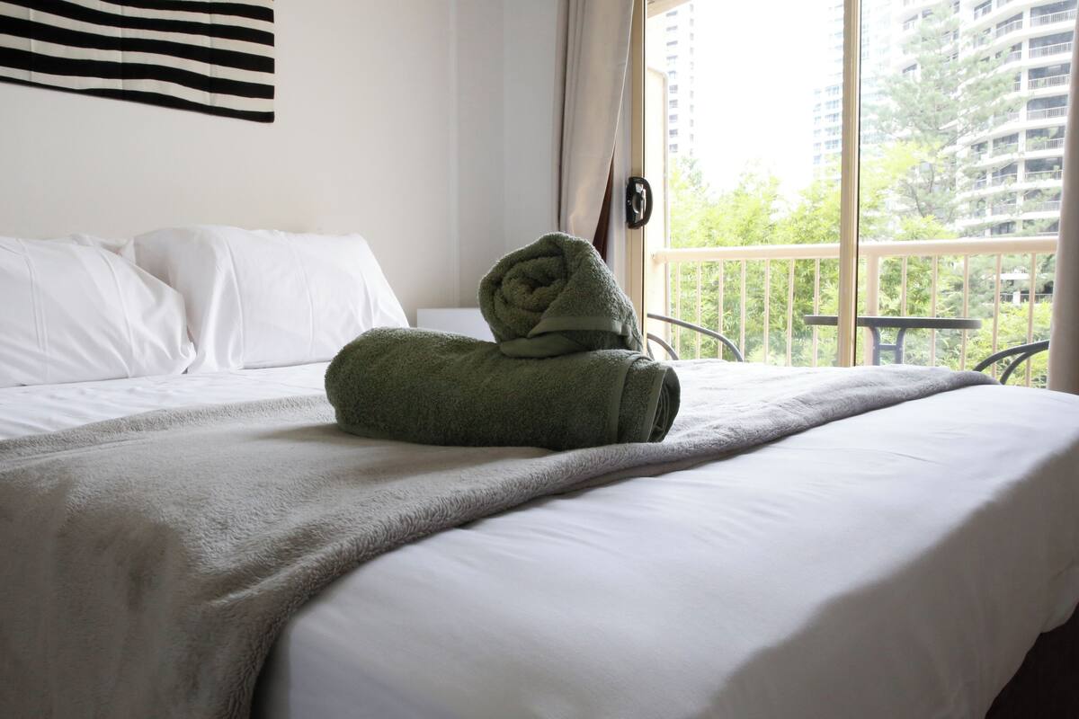 Hotellihuone, jossa on siististi pedattu sänky, parveke, jolta on näkymä vehreään maisemaan ja rakennuksiin, sekä ikkuna, jossa on verhot.