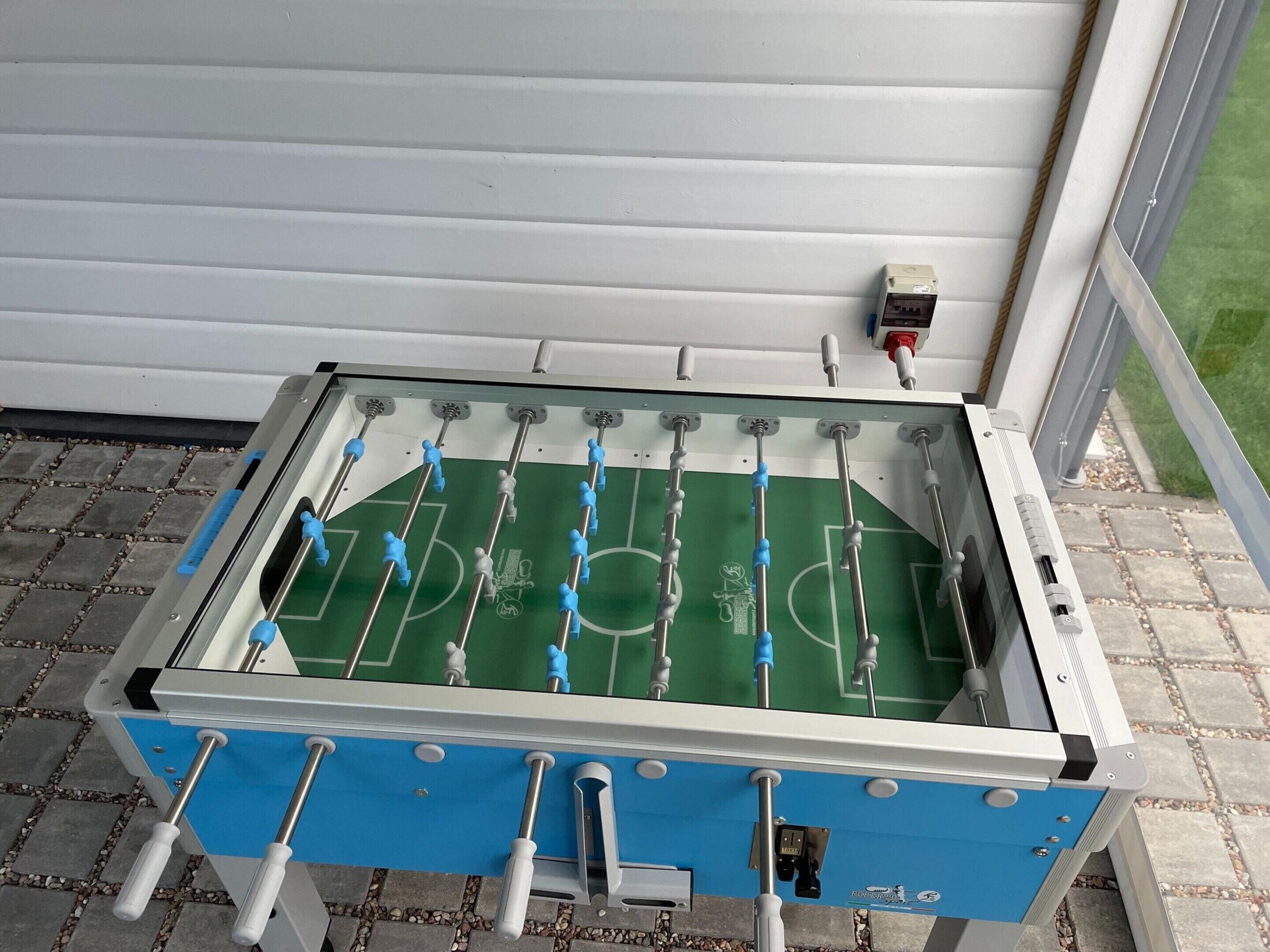 A Foosball table with a blue and white color scheme, located indoors with a window and a wall-mounted electrical outlet visible.